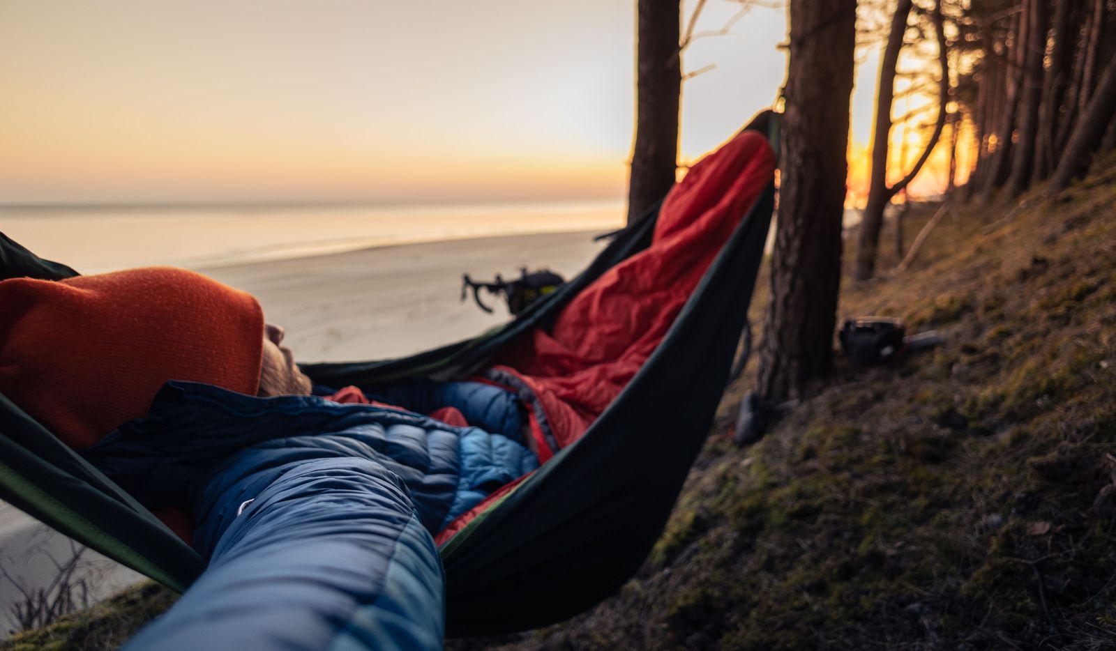 Hammock camper wrapped in a red sleeping bag at a beach sunset