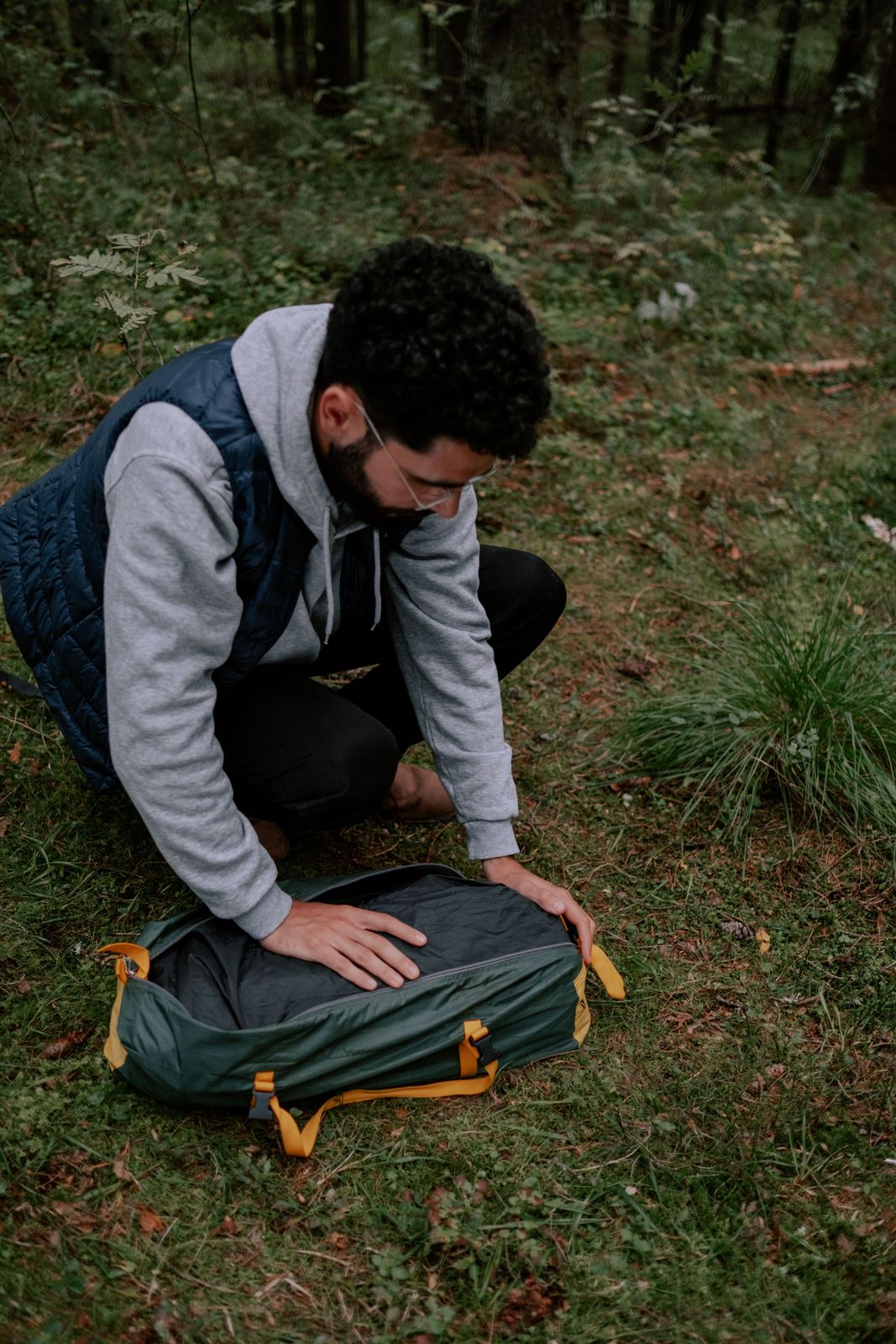 Camper packing a sleeping bag into a green duffel on the forest floor